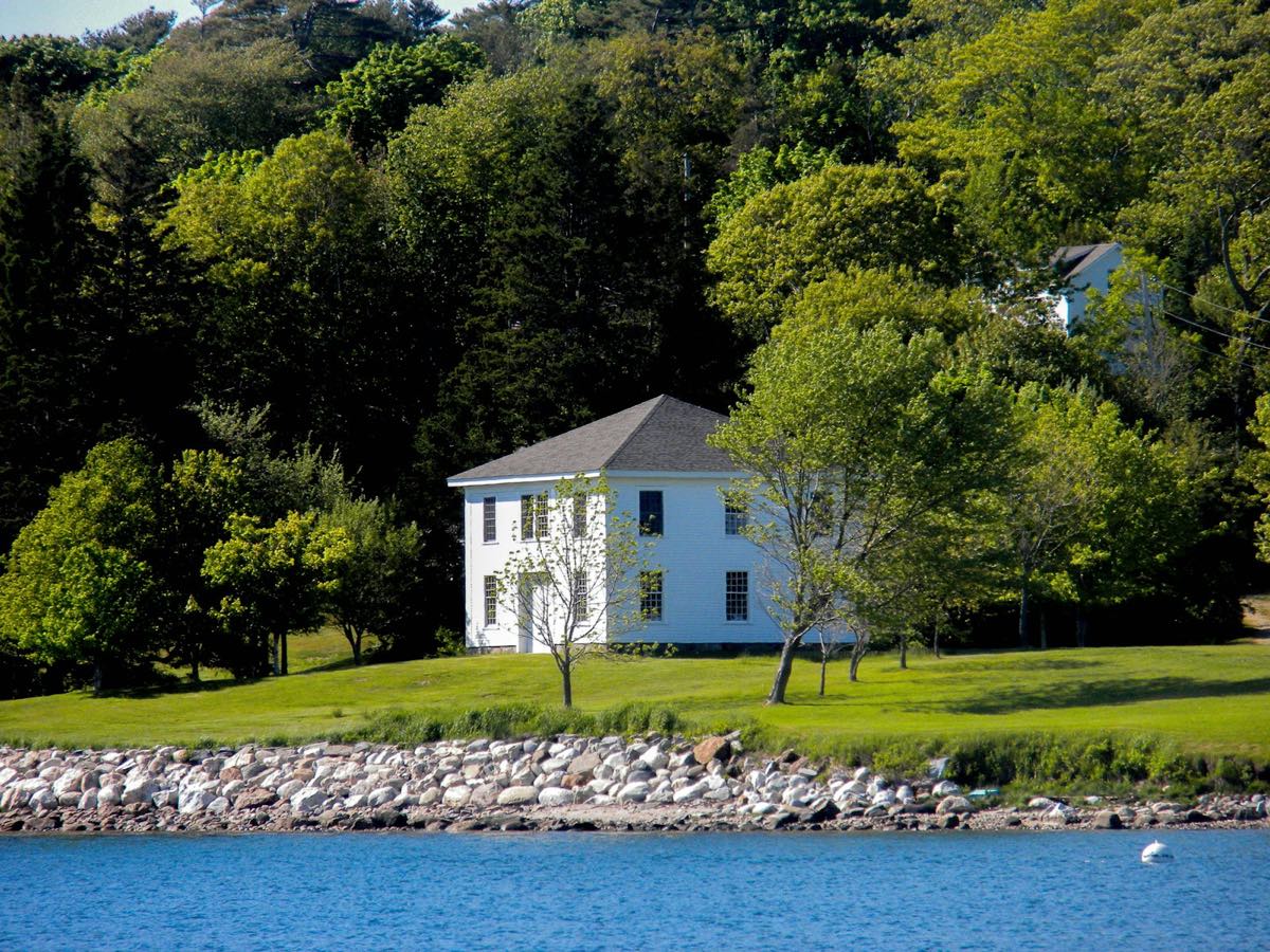 A white two-story house on a lawn above a rocky shore, with trees behind it